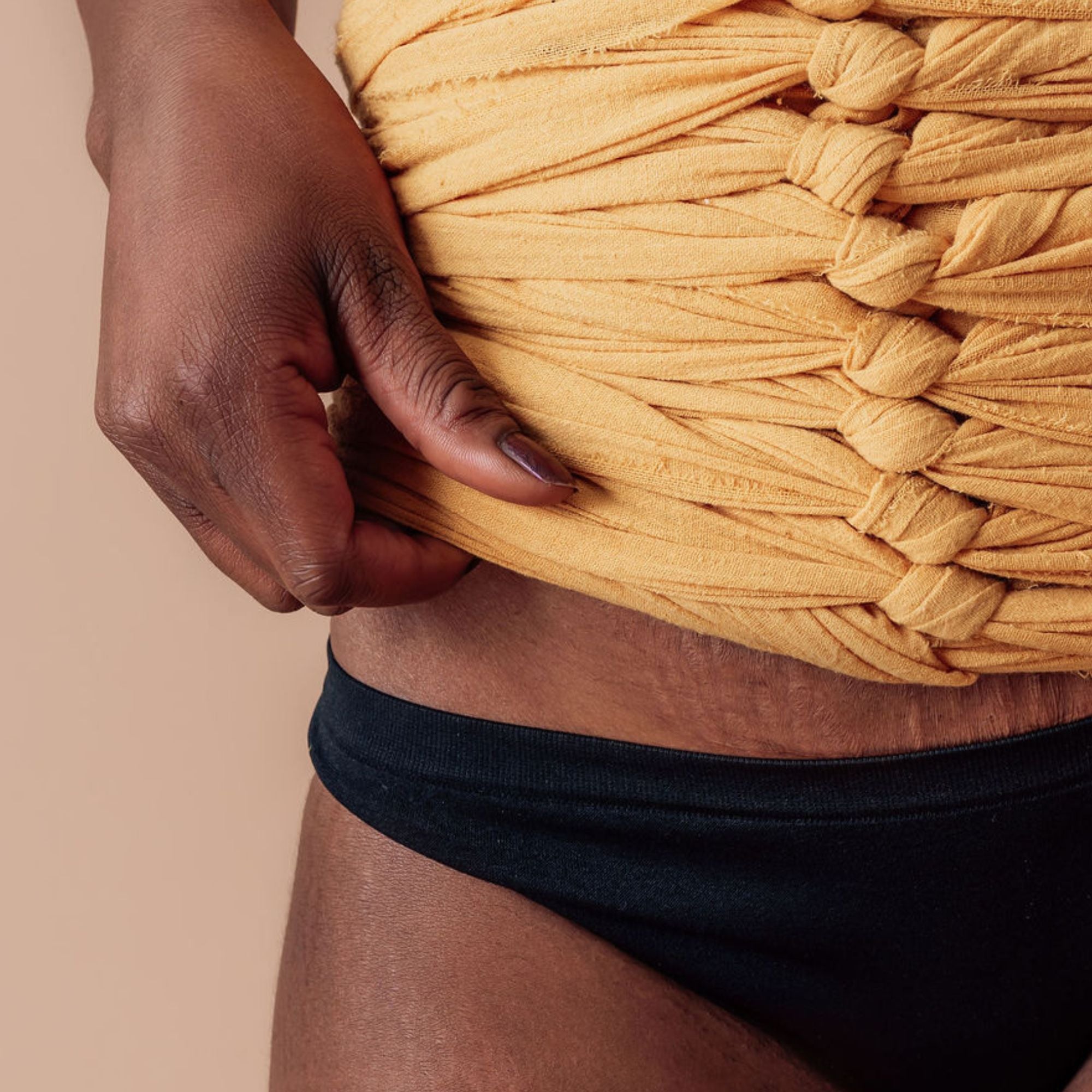 Close-up of a mother adjusting a mustard yellow BelliBind bengkung belly wrap, highlighting its gentle, customized fit over natural postpartum skin and stretch marks.