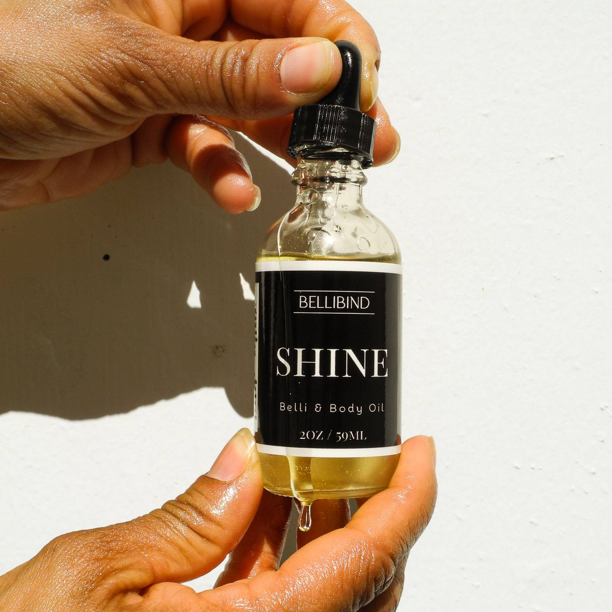 Bellibind Shine Belli and Body Oil held in both hands, a drop of golden castor oil falling from the bottle.